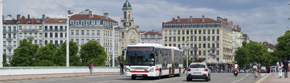 A Keolis bus on a Lyon street, with the city's stone facades and a bridge behind it.